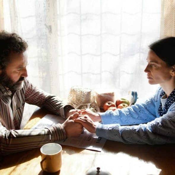 A man and woman sit at a wooden table holding hands in front of a window.
