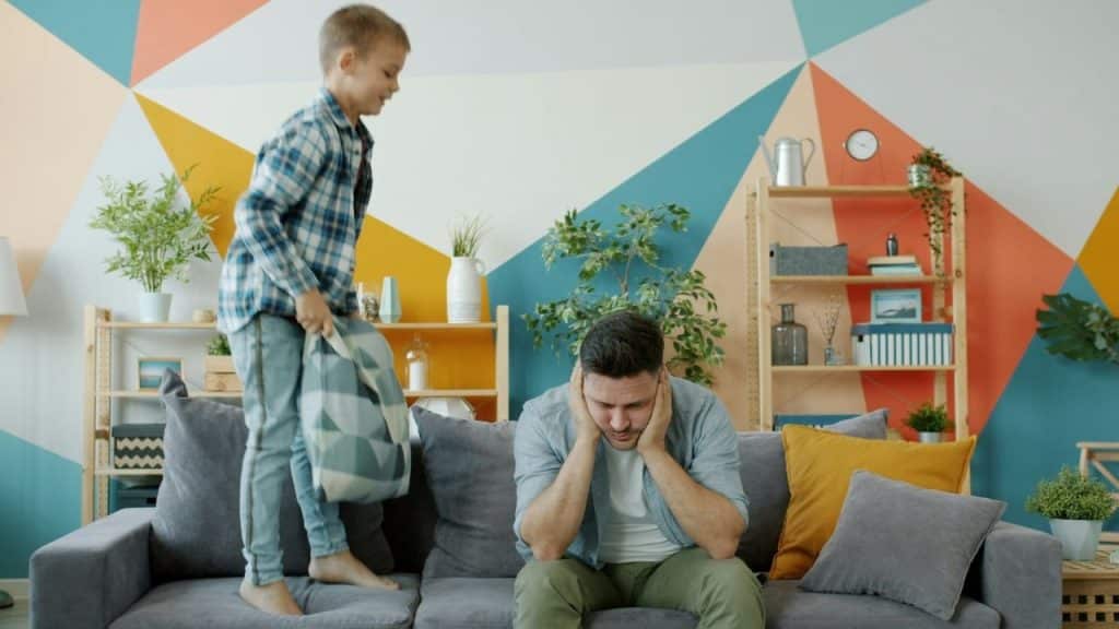 A man sits on a couch covering his ears while a young boy jumps beside him.