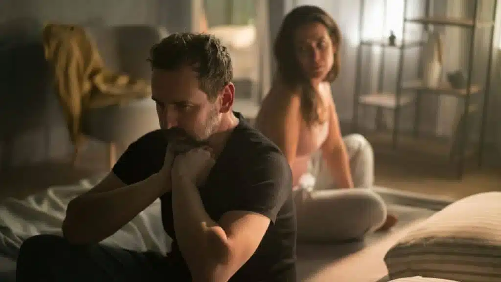 A man and woman sit on a bed in a dimly lit room, facing away.