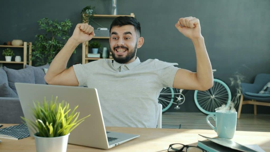 A man sits at a desk in front of a laptop with his arms raised.