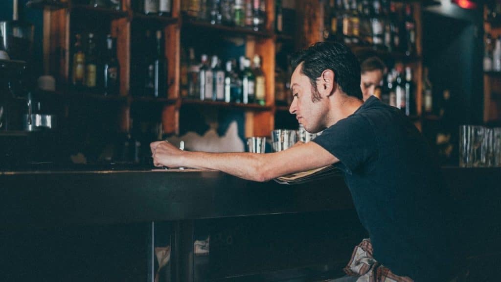 A man leans on a dark bar counter in a room with shelves of bottles.