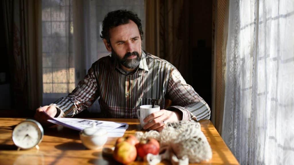 A bearded man sits at a table holding a mug and looking out a window.