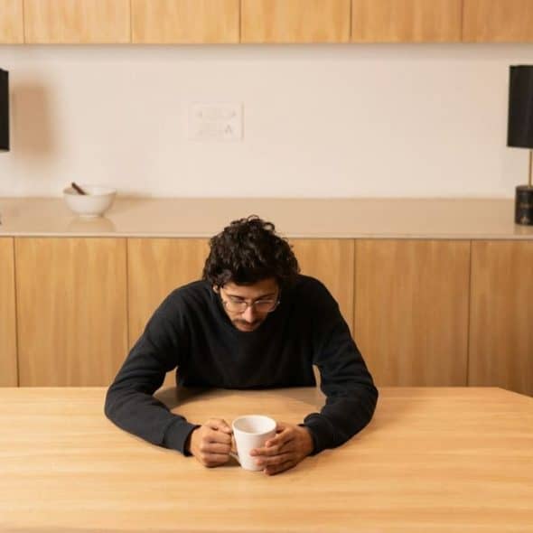 A man with curly hair and glasses looking down at a white mug on a table.