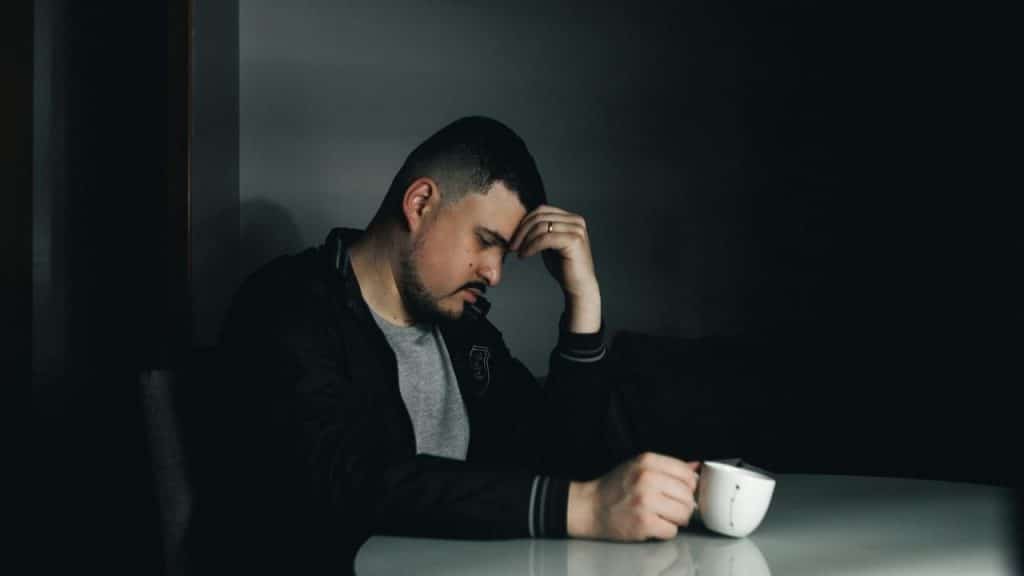 A man in a dark jacket sitting at a table with his hand on his forehead.