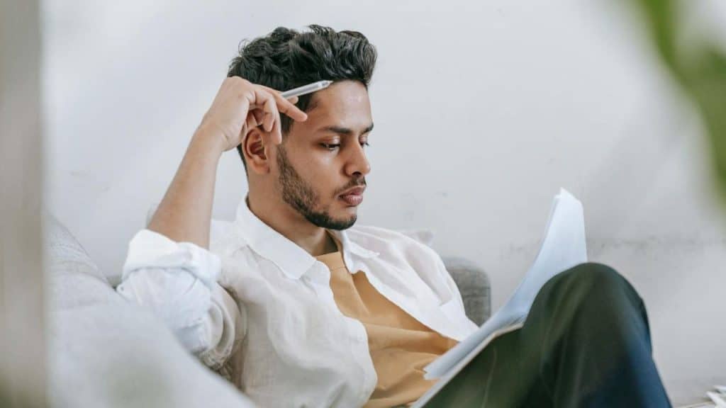 A man sitting on a couch holding a pen to his head while reading paper documents.