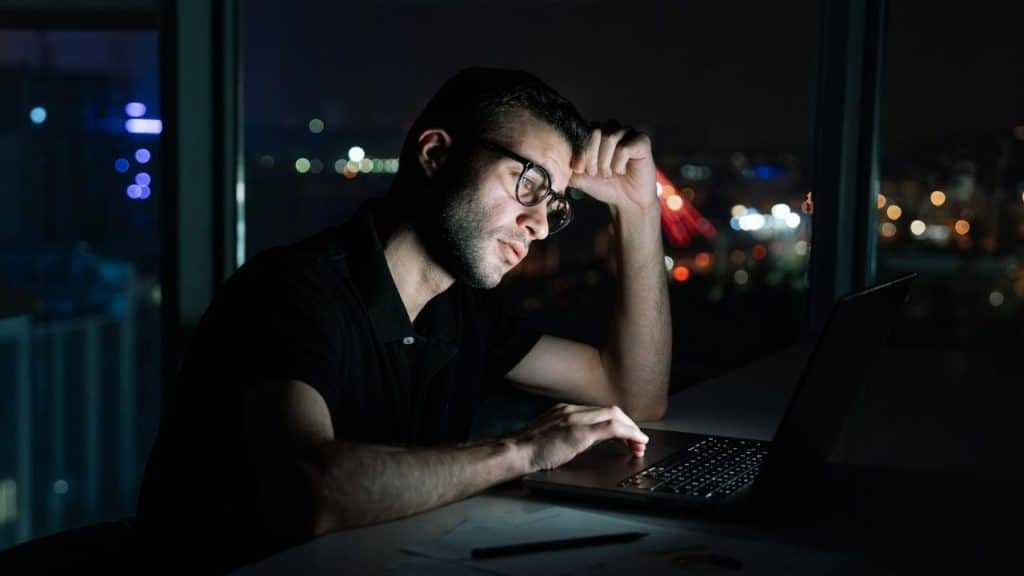 A man with glasses working on a laptop at night with city lights in the background.