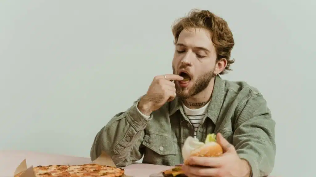 A man in a green corduroy shirt eating a burger and pizza at a pink table.