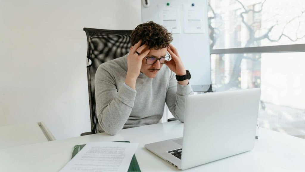 A man with glasses and curly hair holding his head while sitting at a laptop computer.