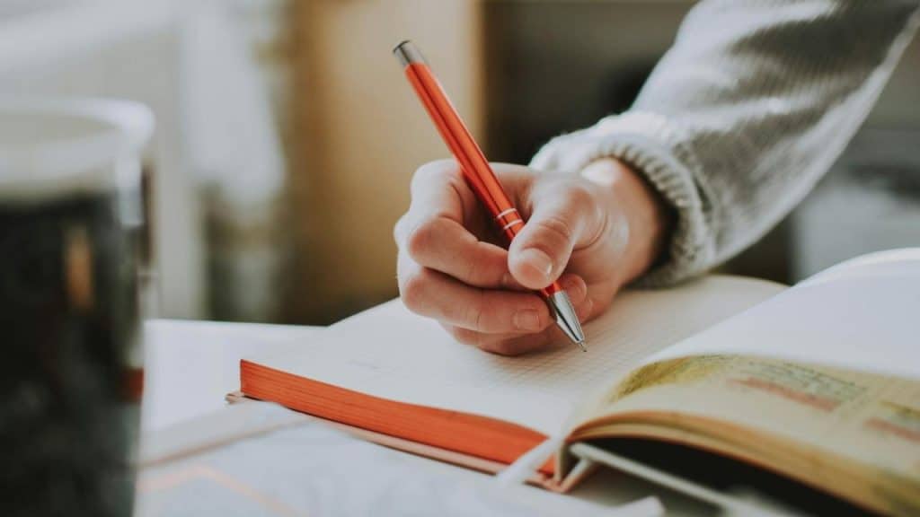 A close-up shot of a person's hand writing in a notebook with an orange-colored pen.