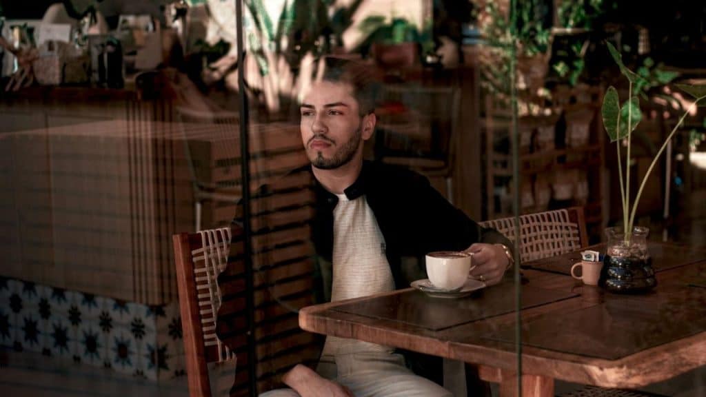 A man with a beard sitting at a wooden table holding a cup behind a glass window.