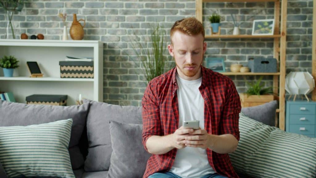 A man in a red plaid shirt sitting on a couch and looking at a smartphone.