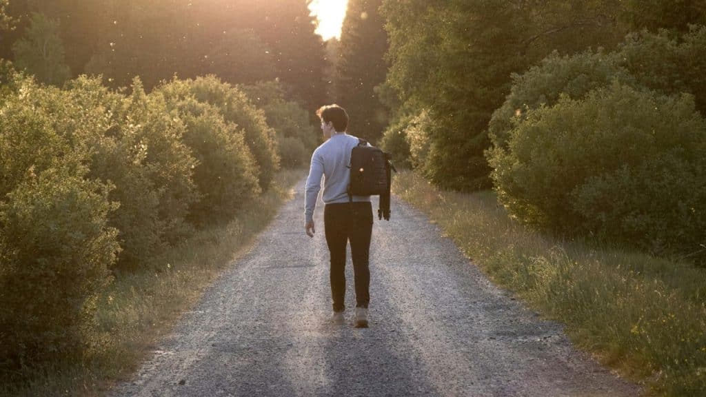 A man with a backpack walking down a dirt path surrounded by trees at sunset.
