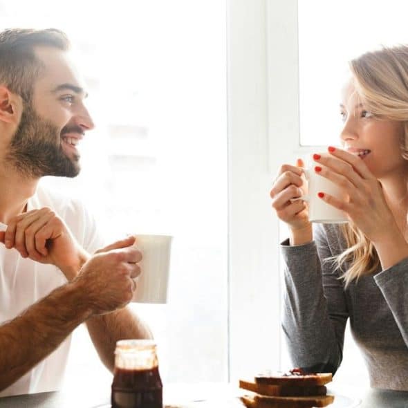 A man and woman sit at a table, smiling and holding mugs near a window.