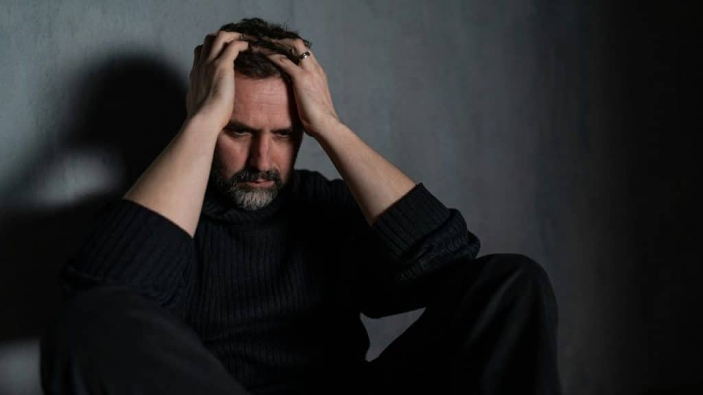 A man with a beard sits against a dark wall with his hands on his head.