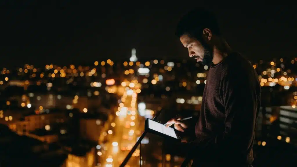 A man on a dark balcony uses a glowing tablet with city lights in the background.