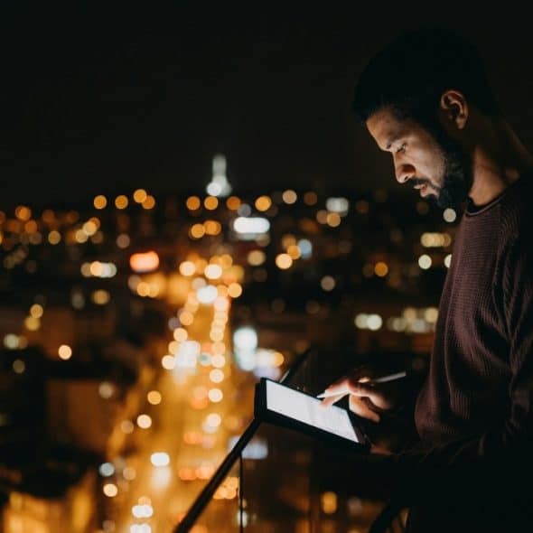 A man on a dark balcony uses a glowing tablet with city lights in the background.