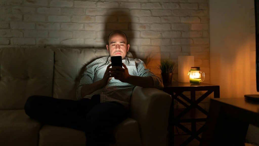 A man sits on a dark sofa in a dim room looking at a bright smartphone screen.