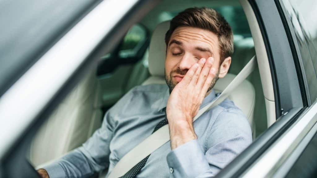 A man in a car rests his head on his hand with his eyes closed tight.