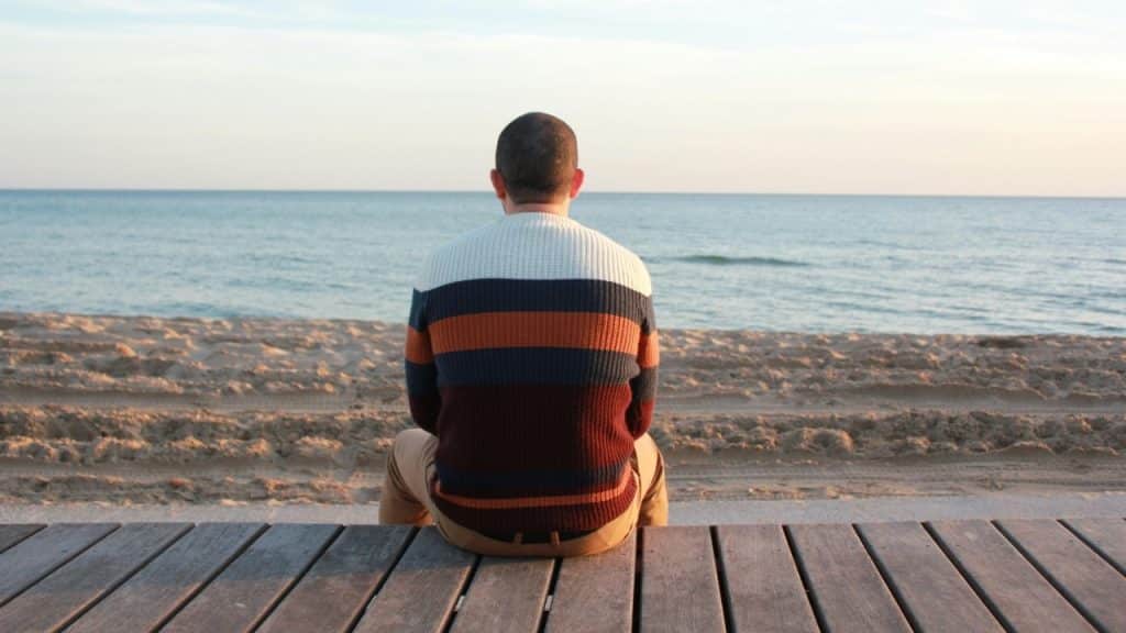 A man in a striped sweater sits on a wooden deck looking out at the ocean.