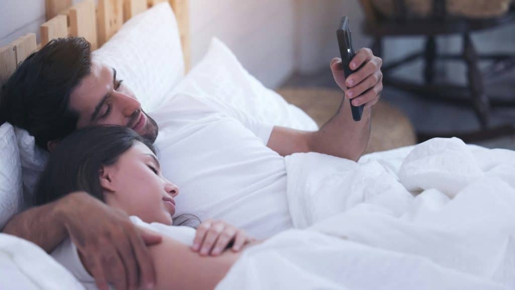 A man lies in bed looking at his phone while a woman sleeps beside him.