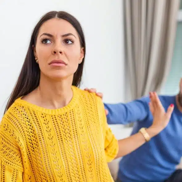 A woman in a yellow sweater looks away while a man reaches toward her shoulder.