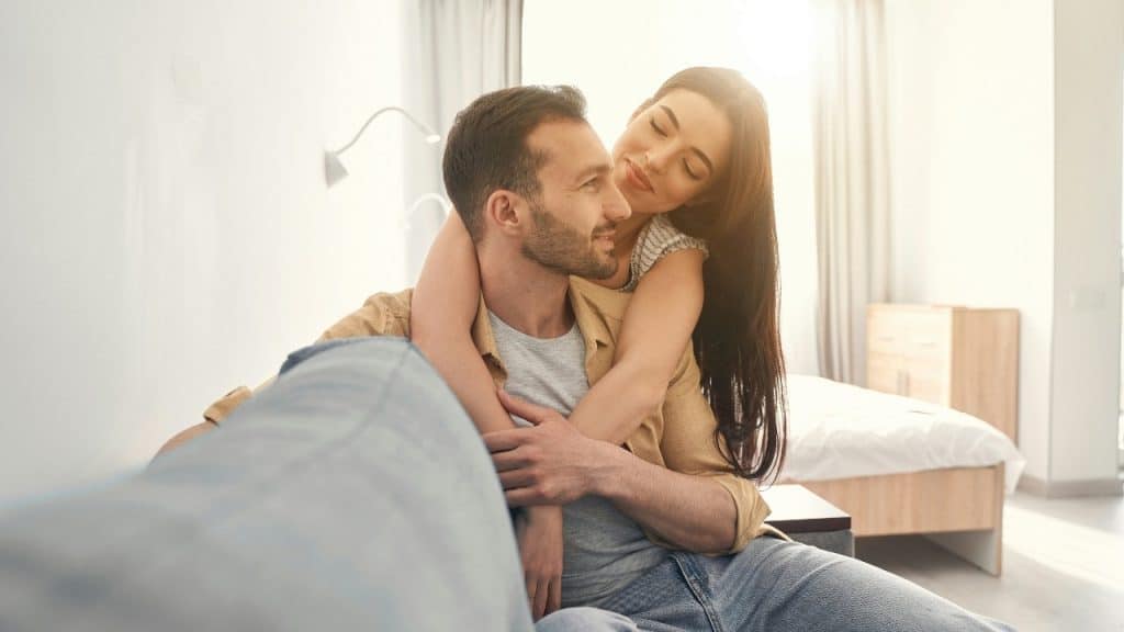 A woman smiles while wrapping her arms around a man sitting on a light-colored sofa.