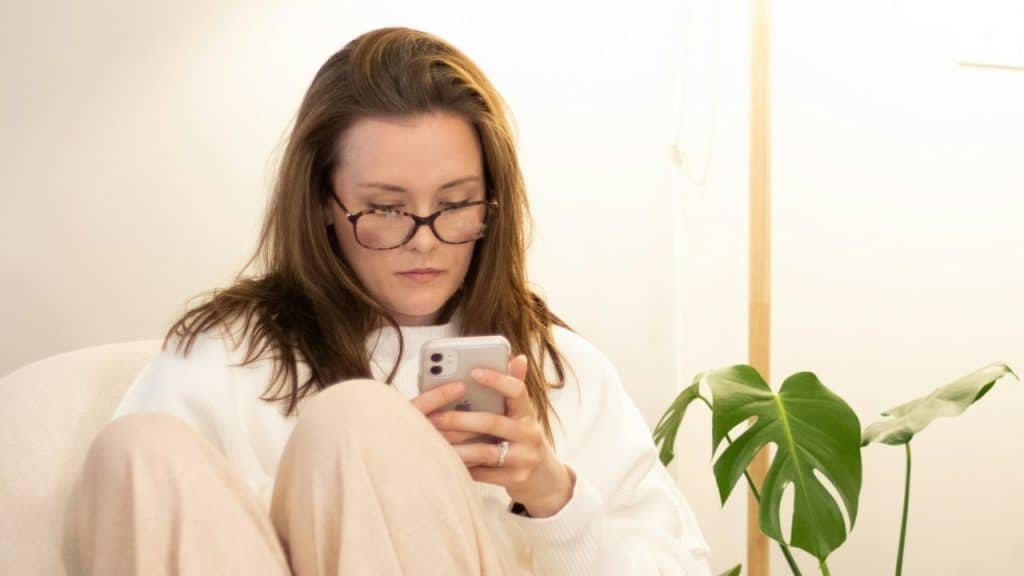 A woman wearing glasses sits indoors while looking down at the screen of her mobile phone.