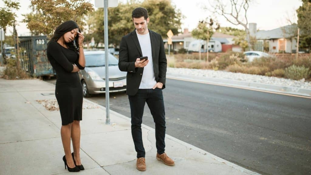 A woman looks down while a man in a blazer stands nearby looking at his phone.