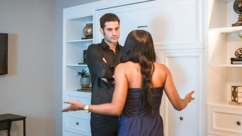 A woman with her back to the camera gestures toward a man with crossed arms.