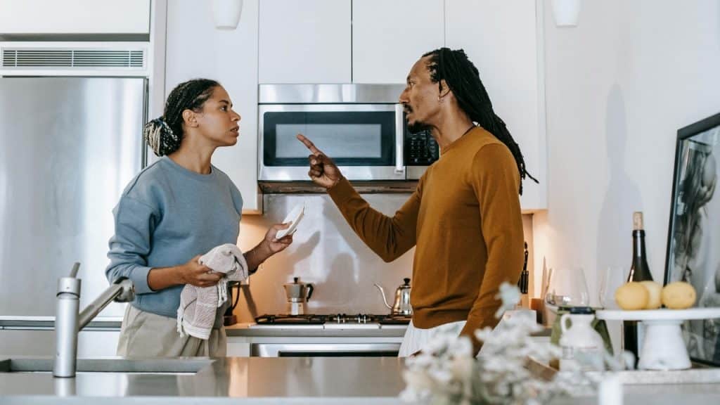 A man points a finger at a woman holding a dish and towel in a kitchen.