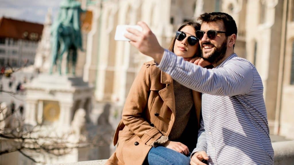 A man and woman wearing sunglasses take a selfie together in front of a stone building.