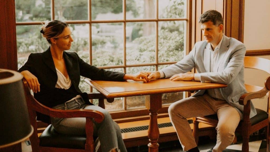 A man and woman sit at a wooden table in a formal room holding each other's hands.