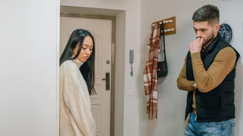 A man and woman stand in a hallway, both looking down with serious facial expressions.