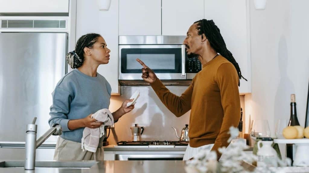A man points his finger while speaking to a woman holding a dish in a kitchen.