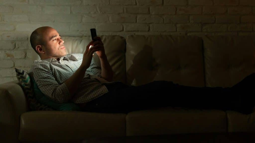 A man reclines on a leather sofa in a dark room while looking at a smartphone.