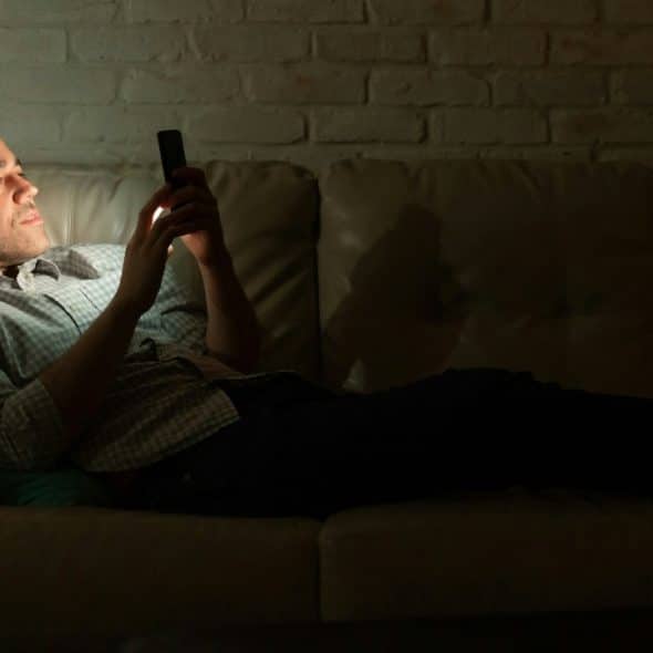 A man reclines on a leather sofa in a dark room while looking at a smartphone.