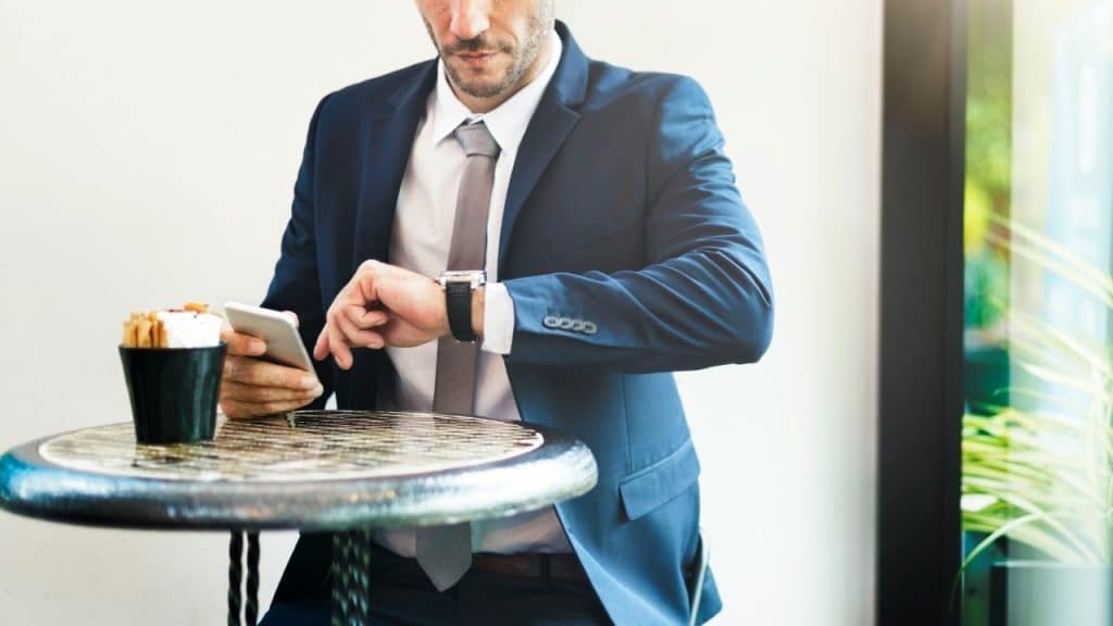 A man in a blue suit sits at a cafe table while checking his wrist watch.