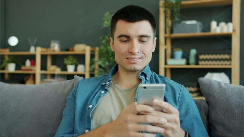 A man in a denim shirt sits on a grey sofa while looking at a smartphone.