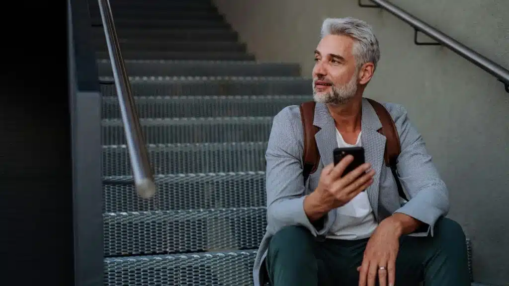 A man with grey hair sits on outdoor metal stairs while holding a mobile phone.