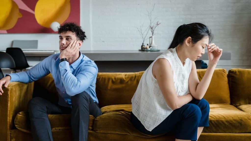 A man and woman sit apart on a mustard-colored couch with their heads turned away.
