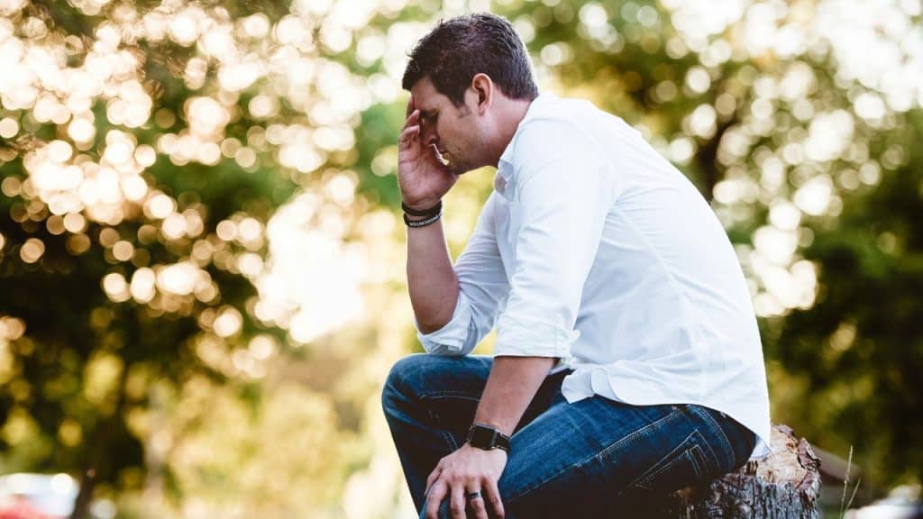 A man in a white shirt and jeans sits outdoors with his hand on his forehead.