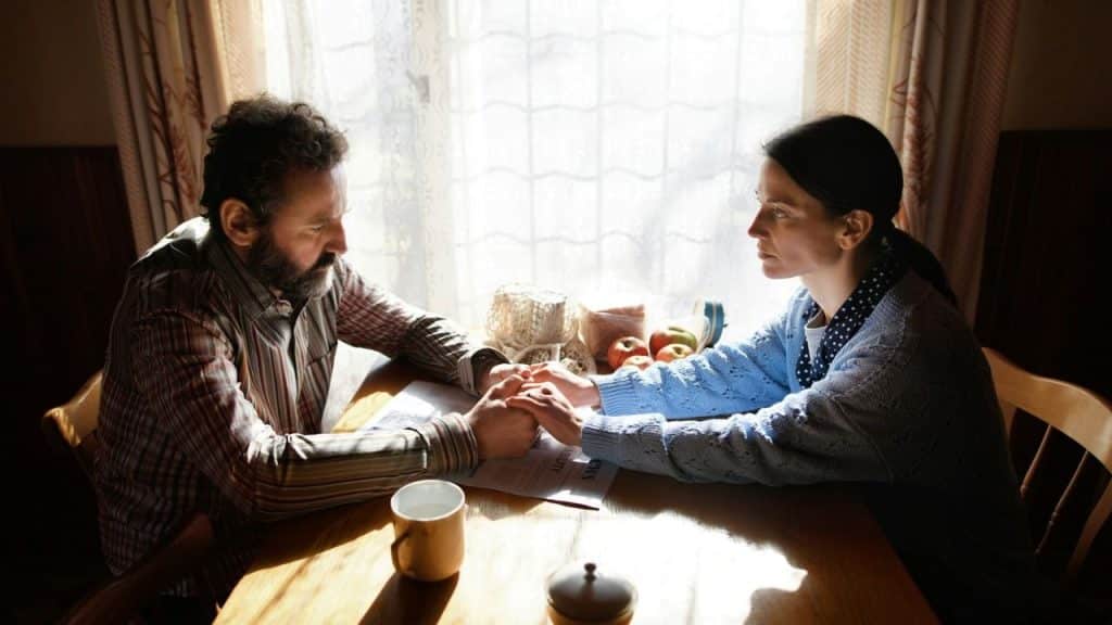 A man and woman sit at a wooden table, holding hands while looking at each other