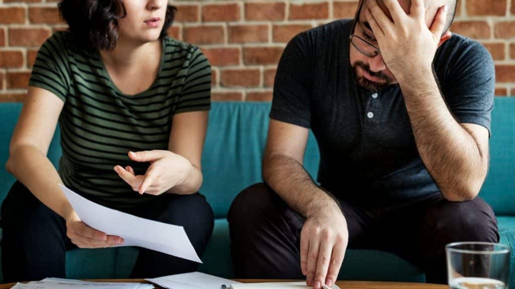 A woman holds a paper and gestures while a man sits beside her with eyes closed.