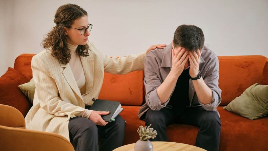 A woman in a blazer rests her hand on a man’s shoulder while he hides his face.