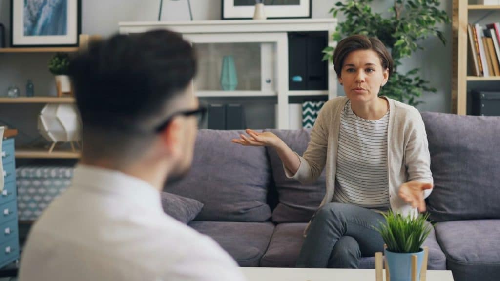 A woman sits on a purple couch and gestures with both hands while speaking to a man.