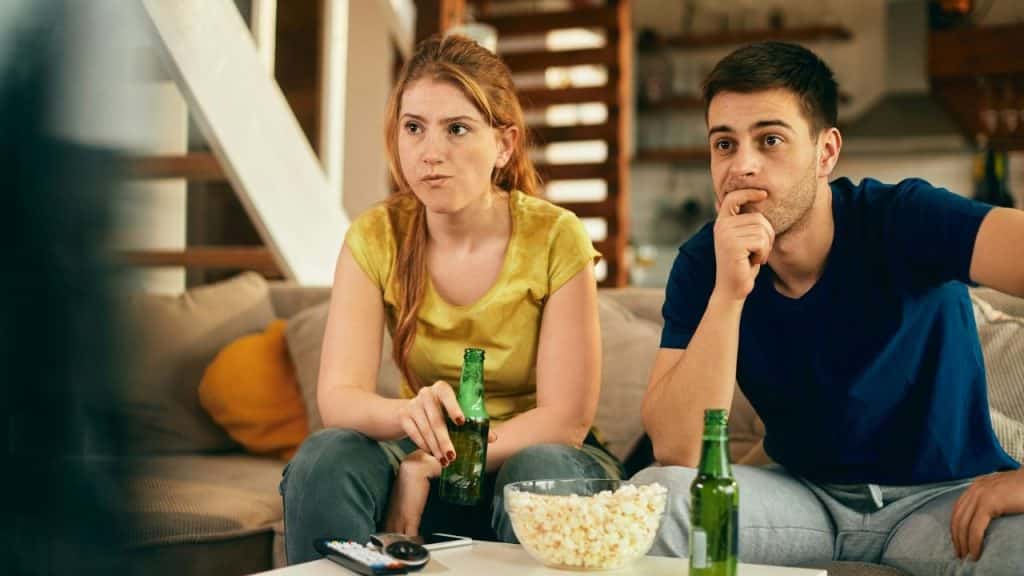 A woman and man sit on a couch, each holding a green glass bottle while watching television.