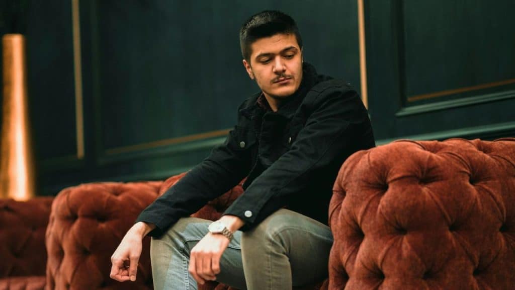 A man in a black jacket sits on a tufted reddish-brown sofa, looking downward.