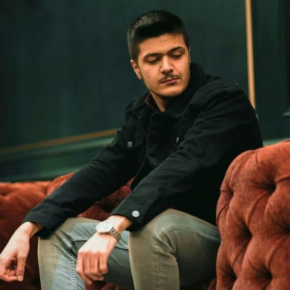 A man in a black jacket sits on a tufted reddish-brown sofa, looking downward.