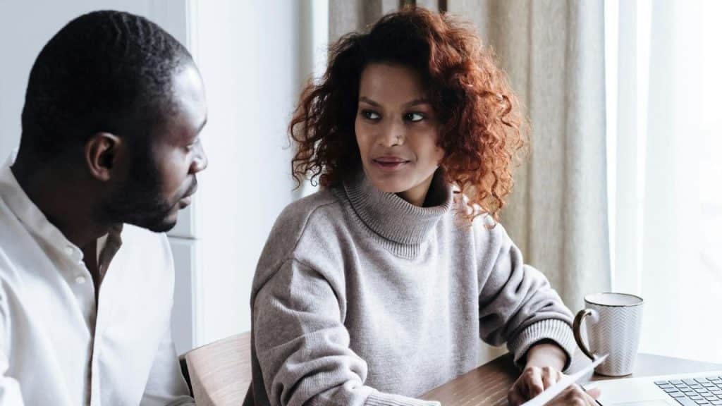 A woman with curly hair looks at a man while sitting at a desk with a laptop.