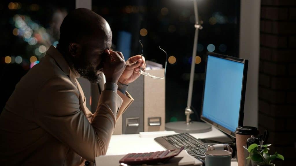 A man in a suit rubs his eyes while sitting at a desk at night.